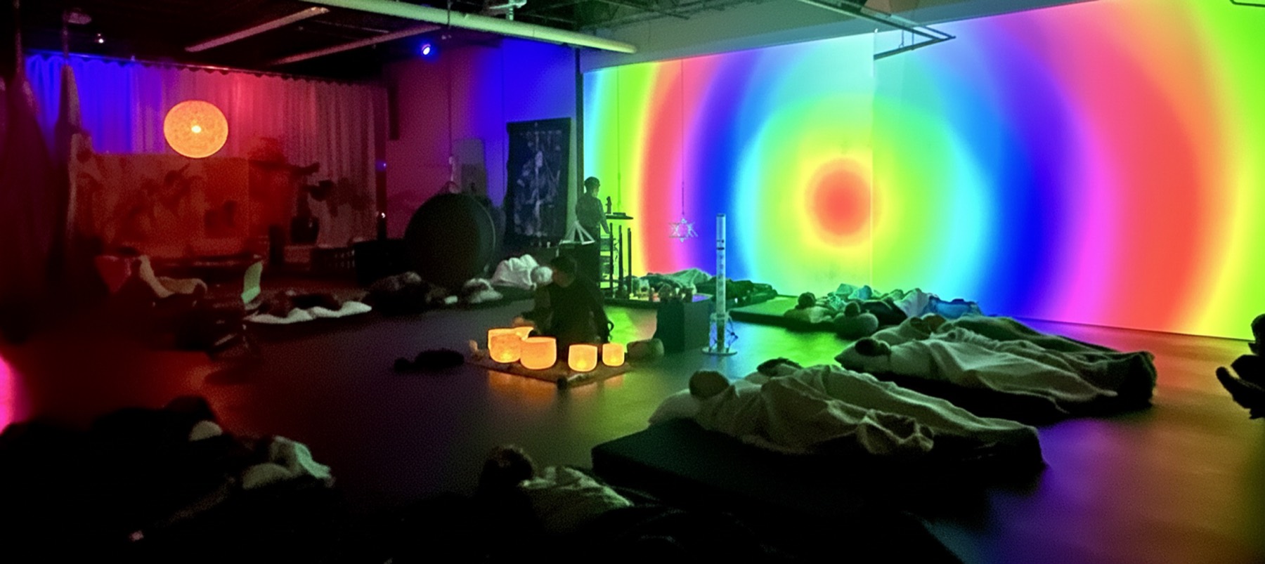 Concentric rainbow projection on the wall with glowing sound bowls and resting guests